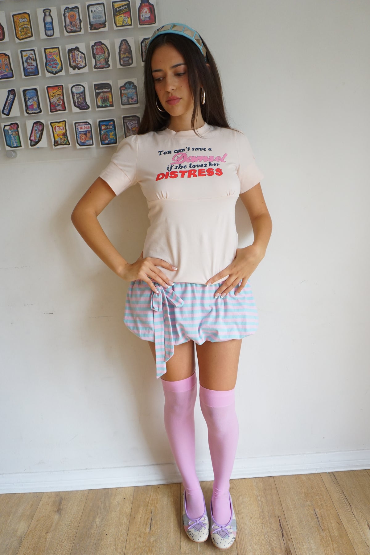 girl wearing peach pink t shirt with buttons on the sleeve and a graphic that says "you can’t save a damsel if she loves her distress"
