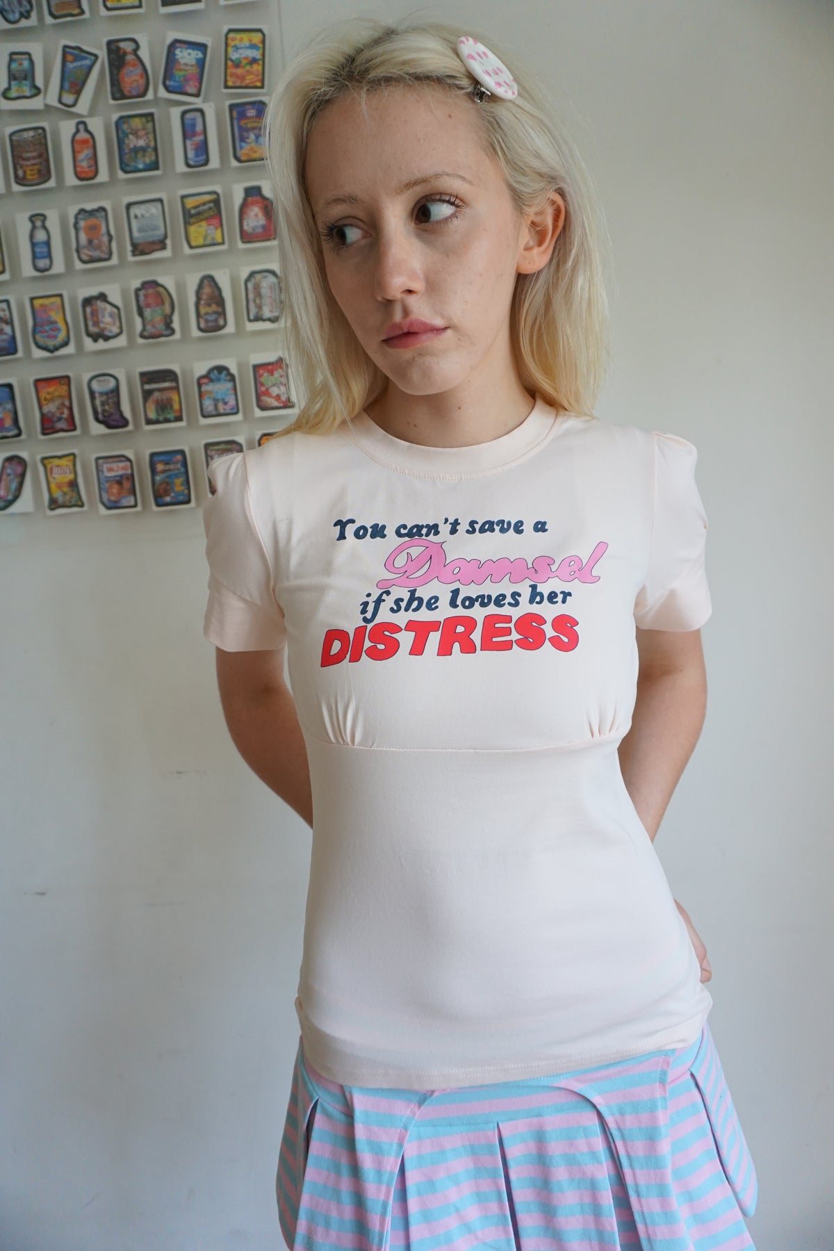 girl wearing peach pink t shirt with buttons on the sleeve and a graphic that says "you can’t save a damsel if she loves her distress"
